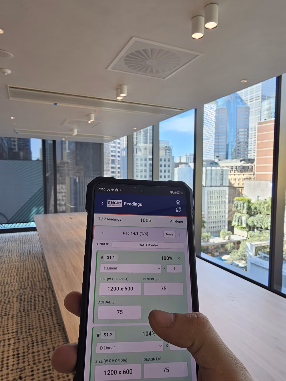 ENGIC's report app on site showing completed air balancing readings, held in hand in a Sydney CBD office with the skyline behind